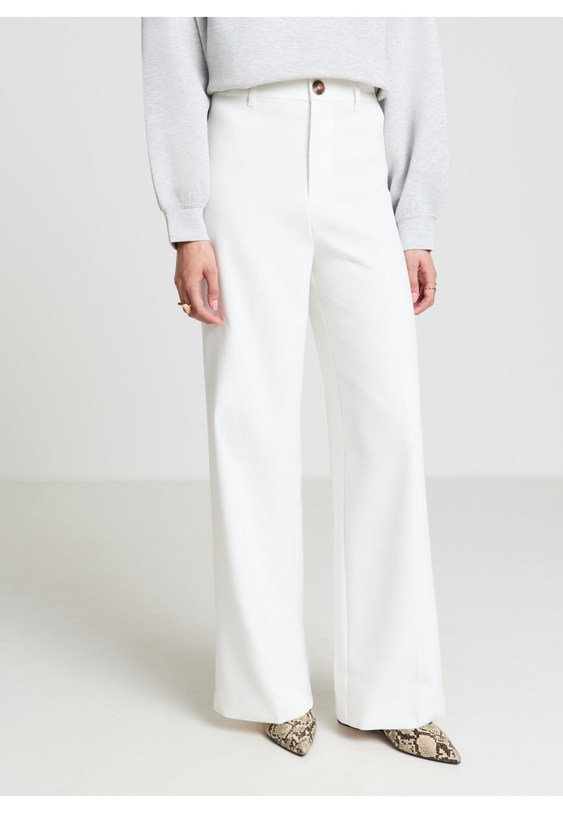 Pantaloni bianchi a gamba larga con vita alta, dotati di tasche frontali e chiusura con un solo bottone marrone, abbinati a un maglione grigio e décolleté con motivo pattern.