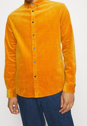 Person trägt ein hell oranges Kordhemd mit Kragen und blauer Jeans, steht vor einem hellen Hintergrund.