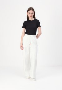 Camiseta negra de manga corta combinada con pantalones anchos blancos de tiro alto con cordón y costuras visibles. La modelo está de pie sobre un fondo liso.
