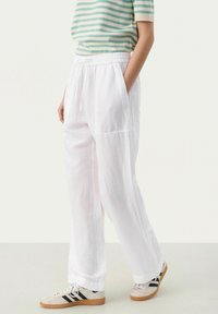 Pantalons en lin blancs et larges avec une taille élastique et des poches latérales. Un mannequin porte un top à rayures vertes et beiges et des baskets.