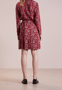 Czerwona sukienka w kwiatowy wzór z długimi rękawami, ściąganym w pasie, plisowanym dołem oraz czarnymi butami z ćwiekami, ukazująca widok z tyłu.
