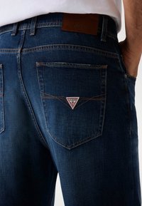 Tamsiai mėlynos džinsinės kelnės su odiniu ženkleliu, pažymėtu "GUESS". Turi dvi užpakalines kišenes su raudonais ir baltais trikampio formos logotipo akcentais.