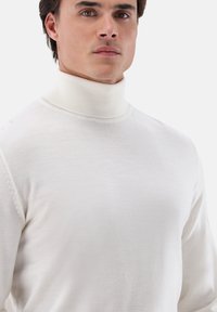 Junger Mann in einem enganliegenden weißen Rollkragenpullover, von der Brust aufwärts vor einem schlichten hellen Hintergrund dargestellt.