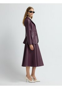 Blazer in pelle bordeaux con due tasche, abbinato a una gonna A-line coordinata. Il modello indossa occhiali da sole neri e scarpe con tacco bianche.