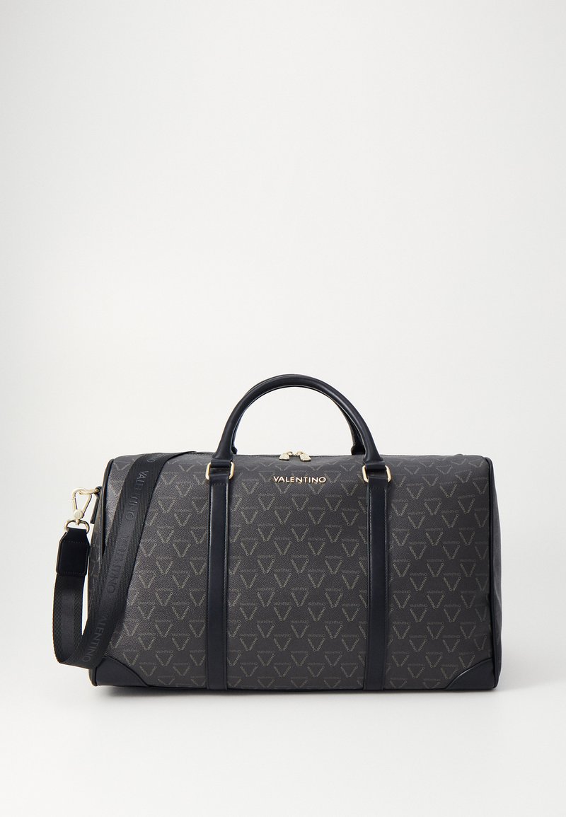 Μαύρη καπιτονέ τσάντα ταξιδιού με τριγωνικό σχέδιο σε τονική απόχρωση, χρυσές λεπτομέρειες, διπλές λαβές και αποσπώμενη ιμάντα ώμου.