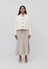 Vrouw met lang zwart haar, gekleed in een crèmekleurige jasje met knopen, een beige midi-jurk en witte hakken met ruffles, staande tegen een grijze achtergrond.