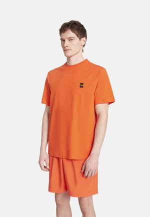 Uomo che indossa una maglietta arancione brillante a maniche corte e pantaloncini coordinati, in piedi davanti a uno sfondo bianco semplice.