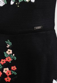 Svart texturerad tyg med röda och rosa broderade blommor och gröna blad, med en liten silverfärgad "GUESS" märkesskylt.