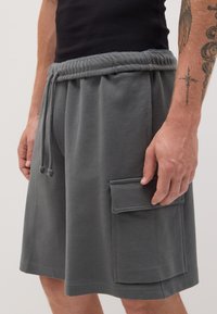 Pantaloni corti in cotone grigio misto con vita elastica, coulisse e una tasca laterale a toppa. Il tessuto ha una consistenza liscia.