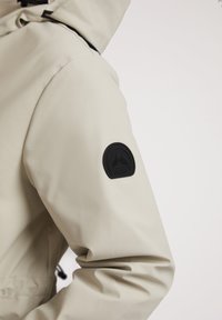 Lichtbeige waterdichte jas met een capuchon, elastische manchetten en een zwart rubberen logo-opdruk op de linkermouw. Soepele textuur.