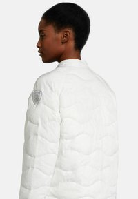 Veste matelassée blanche avec un col arrondi, un motif en vagues texturé et un emblème argenté sur l'épaule gauche.