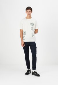 Cremefarbene Baumwoll-T-Shirt mit grünen grafikdesigns, kombiniert mit marineblauen Cargo-Hosen. Schwarze Sneakers mit weißen Streifen und weiße Socken runden den Look ab.