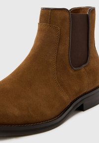 Hnědá semišová Chelsea bota s tmavým elastickým bočním panelem a černou podrážkou, zobrazující detailní šití a hladkou texturu na bílém pozadí.