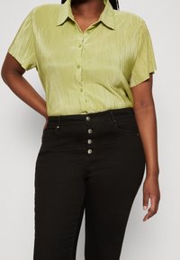 Camisa de manga corta, verde claro, con pliegues y botones en la parte delantera. Combinada con jeans negros de cintura alta que tienen cinco botones metálicos.