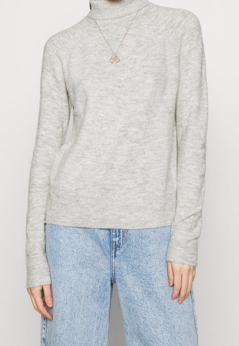 Pull à col roulé gris clair avec une finition texturée, manches raglan et silhouette ajustée, associé à un jean bleu à jambes larges.