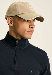Beige beisbola cepure ar izšūtu logo, apvienota ar tumši zilu kapuci, kas ir ar ribotu tekstūru un mazu zilu emblēmu.