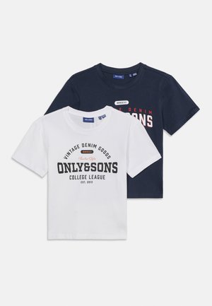 Lächelnder rothaariger Junge, der ein weißes T-Shirt mit Grafik, hellblaue Jeans und weiße Turnschuhe trägt und vor einem weißen Hintergrund steht.