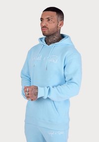 Felpa con cappuccio azzurro chiaro in un tessuto morbido, dotata di tasca frontale e testo del logo bianco. Modello che indossa pantaloni coordinati e accessori.