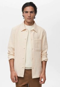 Cream-colored woven shirt jacket with button closure, a chest pocket, and rolled-up sleeves. Textured fabric and relaxed fit.