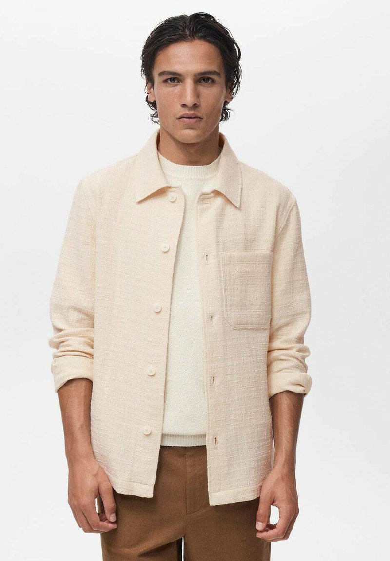 Cream-colored woven shirt jacket with button closure, a chest pocket, and rolled-up sleeves. Textured fabric and relaxed fit.