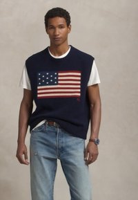 Kék kötött ujjatlan pulóver amerikai zászló mintával, piros, fehér és kék színekben, fehér pólóval és világoskék farmernadrággal viselve.