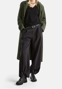 Person som bär en olivgrön kordflöjel lång rock, en svart V-ringad topp, vida svarta byxor med bälte och svarta kraftiga ankelskängor, står med händerna i fickorna.