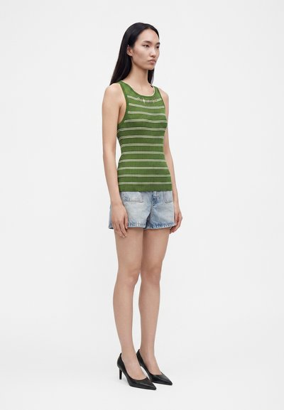 Femme aux longs cheveux noirs porte un haut sans manches rayé vert, un short en jean bleu clair et des talons hauts noirs, debout sur un fond blanc.