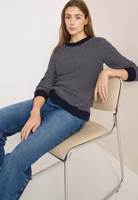 Maglione a maglia blu navy con un motivo testurizzato, polsini e orlo a coste, abbinato a jeans di denim blu, seduto su una sedia in cromo.