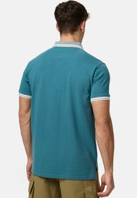 Teal polo-shirt med korte ærmer og hvide striber ved ærmerne og kraven; lavet af blødt stof; bagfra ses et enkelt design.
