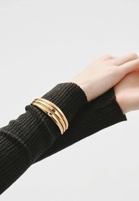 Brazaletes de color dorado con un acabado liso, adornados con piedras de colores, llevados sobre una manga de punto negra. Las bandas están apiladas de cerca.