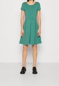 Teal kortärmad klänning med åtsittande överdel och utsvängd kjol, tillverkad av mjukt material, med subtila veck. Matchar med svarta loafers.