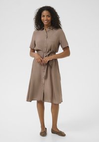 Robe marron, longueur genoux, à boutons avec des manches courtes, taille ceinturée et texture lisse. Portée avec des chaussures plates marron.