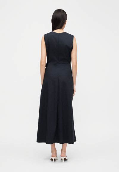 Femme aux longs cheveux noirs portant une robe midi bleu marine sans manches et des chaussures blanches à talons hauts, de dos sur un fond blanc uni.