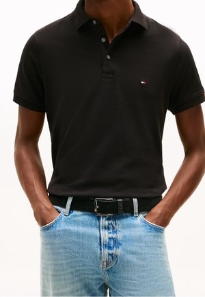 Homme portant un polo noir et un jean bleu clair, les mains dans les poches, ceinture noire avec boucle argentée visible.