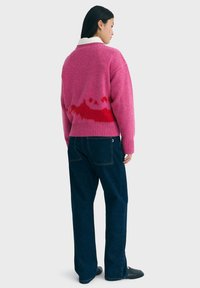 Kvinde iført en pink og rød mønstret sweater over en hvid skjorte med krave, mørkeblå jeans og sorte slip-on sko, stående på siden.