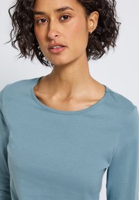 Mladá žena s kudrnatými tmavými vlasy, oblečená v světle modré košili s dlouhým rukávem, která se dívá mírně mimo kameru na jednobarevném bílém pozadí.