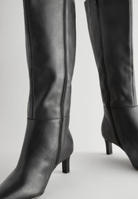 Paire de bottes hautes en cuir noir avec fermetures éclair latérales et talons carrés bas sur un fond neutre.