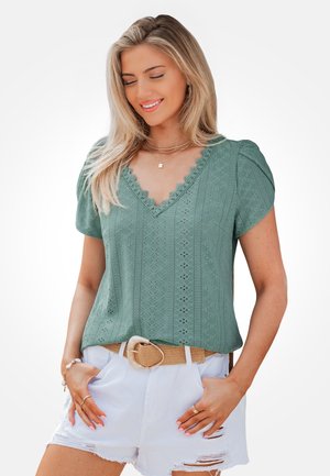 Femme blonde portant un chemisier vert en dentelle avec un décolleté en V, short blanc déchiré avec une ceinture beige, souriant les yeux fermés, les mains dans les poches.