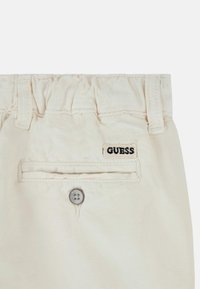 Pantaloni color crema con cintura elasticizzata, tasca posteriore e etichetta logo nera "GUESS". Tessuto morbido con una leggera texture.