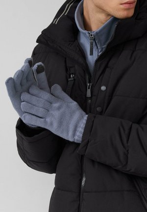 Mann, der eine schwarze Steppjacke und hellblaue Handschuhe mit schwarzen Touchscreen-Spitzen trägt, die Hände zusammengelegt.