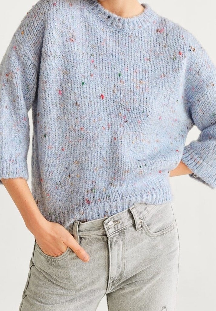 Pull tricot bleu clair avec des taches multicolores, porté avec un jean taille haute gris clair, main dans la poche, coupé à la taille.
