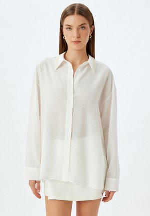 Camicia bianca con bottoni, maniche lunghe, tessuto testurizzato e dettagli decorativi con perline sul colletto. Vestibilità rilassata e orlo curvo nella parte inferiore.
