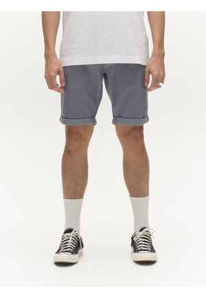 Férfi, aki felhajtott szürke rövidnadrágot, fehér zoknit, fekete alacsony szárú sportcipőt és fehér pólót visel, fehér háttér előtt áll.