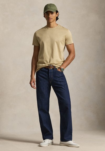 Μπεζ βαμβακερό t-shirt, σκουρό μπλε τζιν παντελόνι, καπέλο baseball ελιάς, λευκά αθλητικά παπούτσια, καφέ ζώνη, casual εφαρμογή, απλός σχεδιασμός, χωρίς σχέδια.