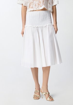 Donna che indossa un crop top in pizzo bianco, una gonna midi bianca a vita alta con cucitura arricciata e sandali beige con tacco alto e borchie dorate.