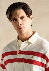 Valge polo särk punaste horisontaalsete triipudega, beeži krae ja väikese kuldse logoga rinnal. Pehme, tekstureeritud kangas.