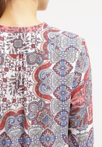Langärmlige Bluse aus leichtem Stoff, mit aufwendigen Paisleymuster in Rot, Blau und Beige, rundem Ausschnitt und Falten-Detail.