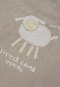 Weißes Cartoon-Lamm mit gelbem Gesicht und Beinen auf beigem Stoff neben dem Text "KLEINES LAMM NOPPIES".