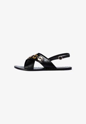 Sandale en cuir noir avec des lanières croisées et des accents en anneau doré. Elle dispose d'une semelle plate avec de petites perforations pour la texture.