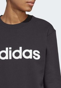 Svart sweatshirt med bred rund halsringning och en framträdande vit "adidas"-logga, tillverkad av mjukt bomullsmaterial.